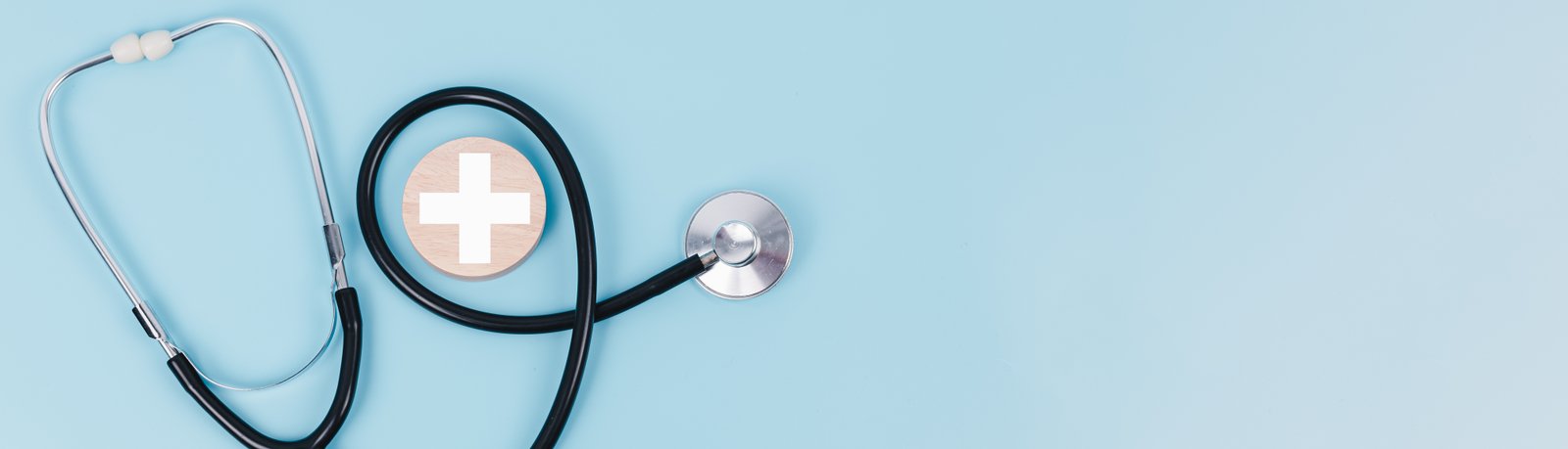 A black medical stethoscope arranged next to a round wooden badge with a white medical cross icon on a light blue background