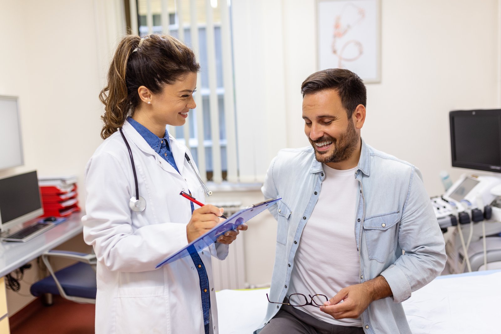 A friendly female doctor in a white coat with a stethoscope holding a clipboard and smiling while talking with a male patient in a medical clinic office