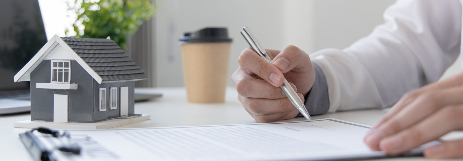 A close-up of a person's hand holding a pen to sign a contract on a desk, next to a small gray architectural model house
